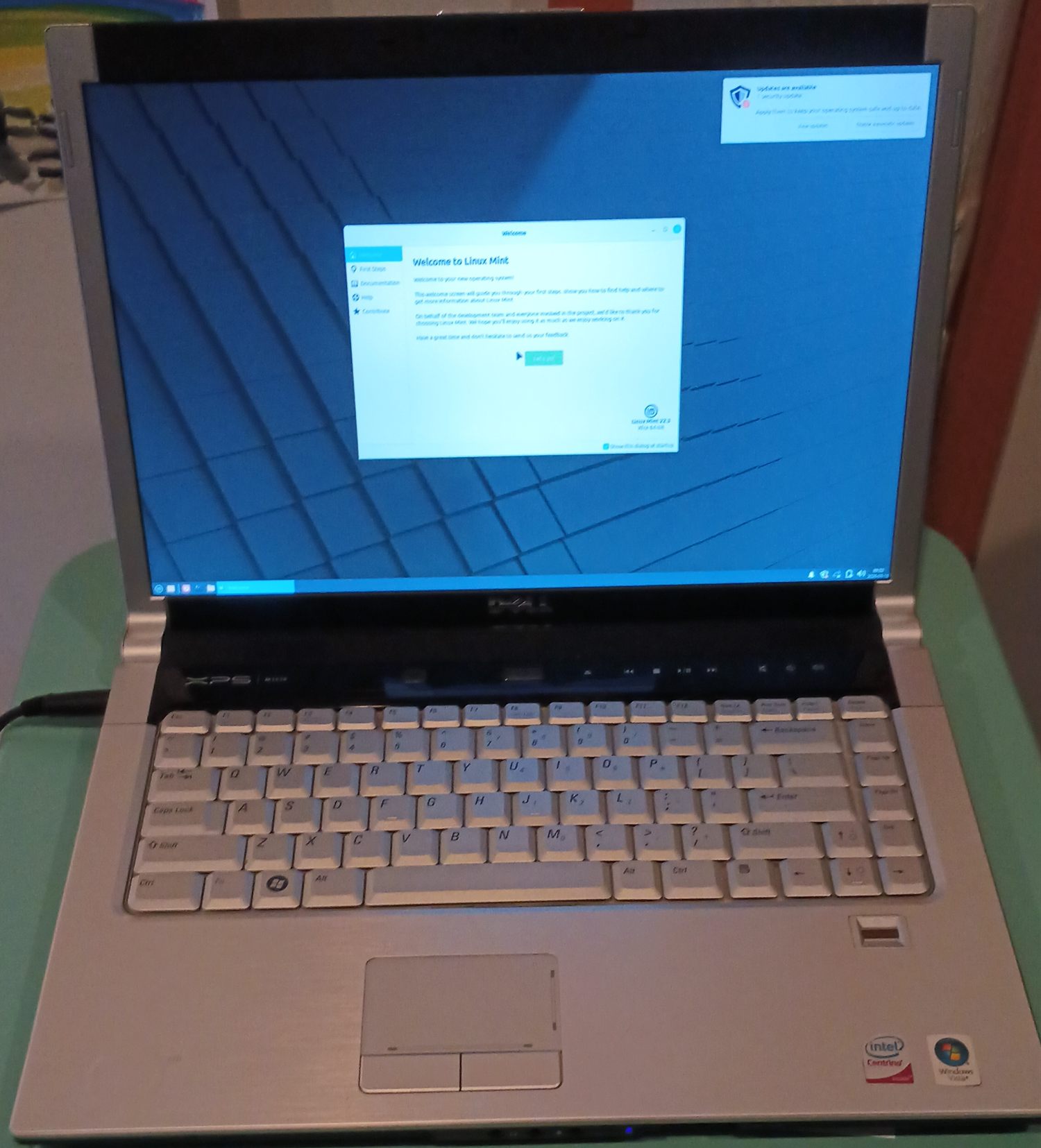 The repaired Dell XPS M1530 open and powered on, running a Linux Mint Xfce desktop from its new SSD. The Welcome to Linux Mint window is open on the blue Mint wallpaper and identifies the release as Linux Mint 22.3 Xfce 64-bit, with a security-update notification in the top right corner. The XPS M1530 badge is visible on the hinge strip above the keyboard, and an Intel Centrino sticker and a Windows Vista sticker are still on the palm rest beside the fingerprint reader.