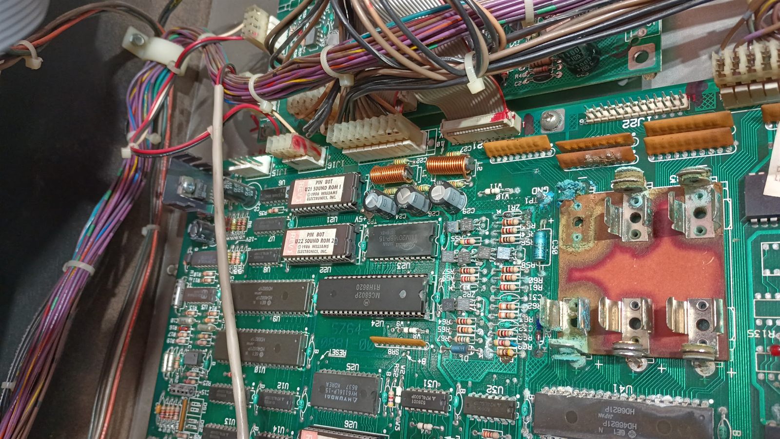 Close-up of the Pinbot System 11 CPU board around the battery holders at B1 to B3. The holder contacts are heavily corroded with blue-green crust, the solder mask has been eaten away over a large area leaving exposed bare board, and the corrosion has spread along the traces toward the surrounding components. The game ROMs labeled PIN BOT U21 SOUND ROM 1 and U22 SOUND ROM 2 are visible to the left, undamaged.