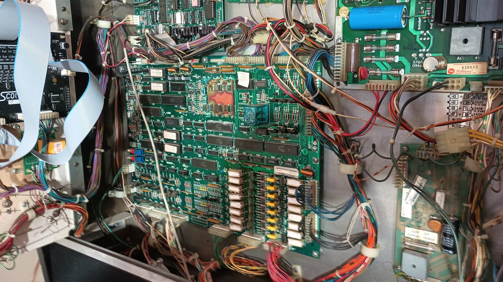 The open backbox of the Pinbot machine showing the full electronics stack: the System 11 CPU and driver board in the center with its rows of power resistors and driver transistors along the bottom edge, the power supply board at upper right with its fuse block and large filter capacitor, the 50 volt flipper power fuse board at lower right, and the wiring harnesses running between them.