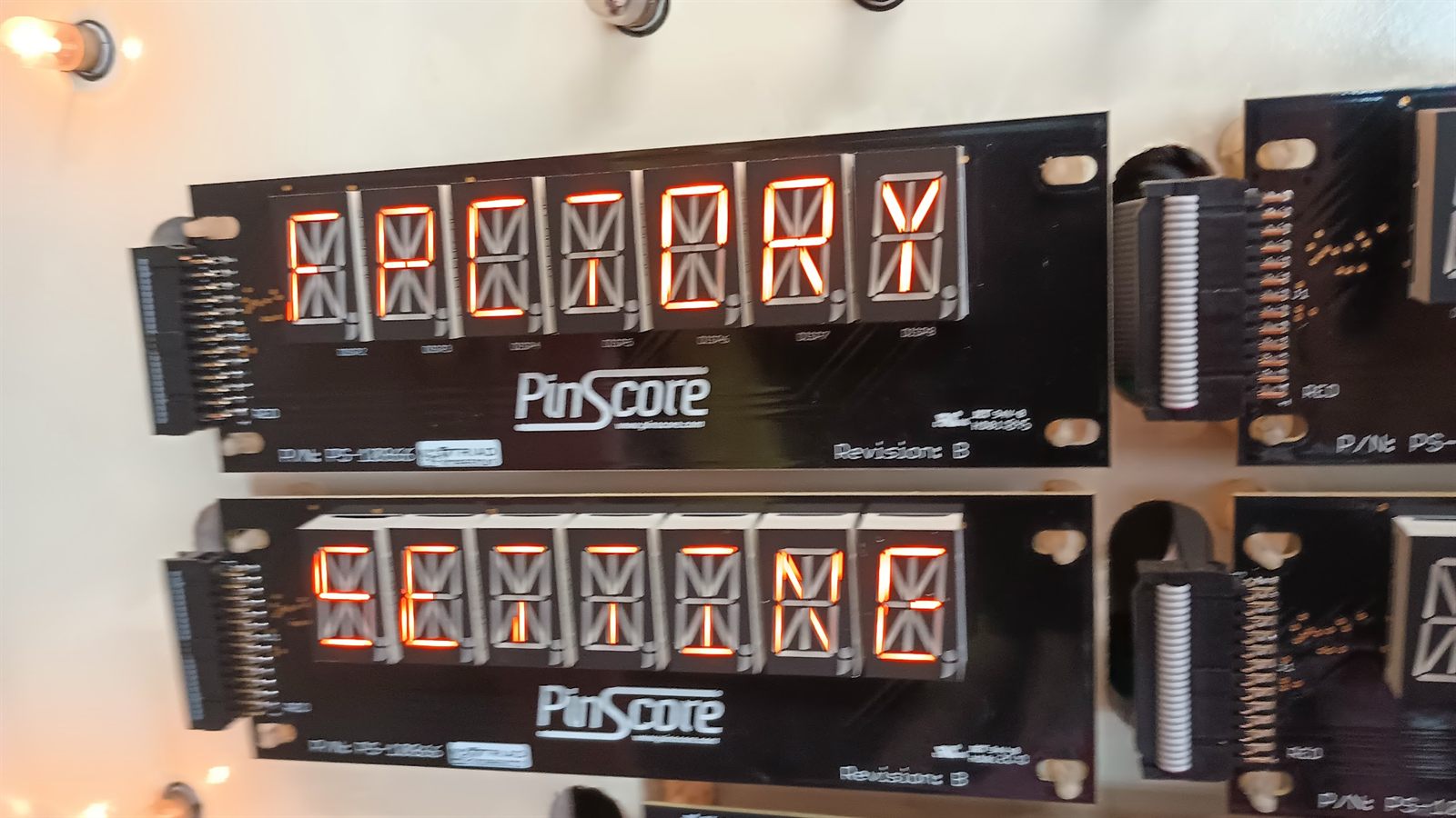 Two alphanumeric display panels fitted in the machine's backbox, lit and reading FACTORY on the upper display and SETTING on the lower one. Both are marked PinScore Revision B.