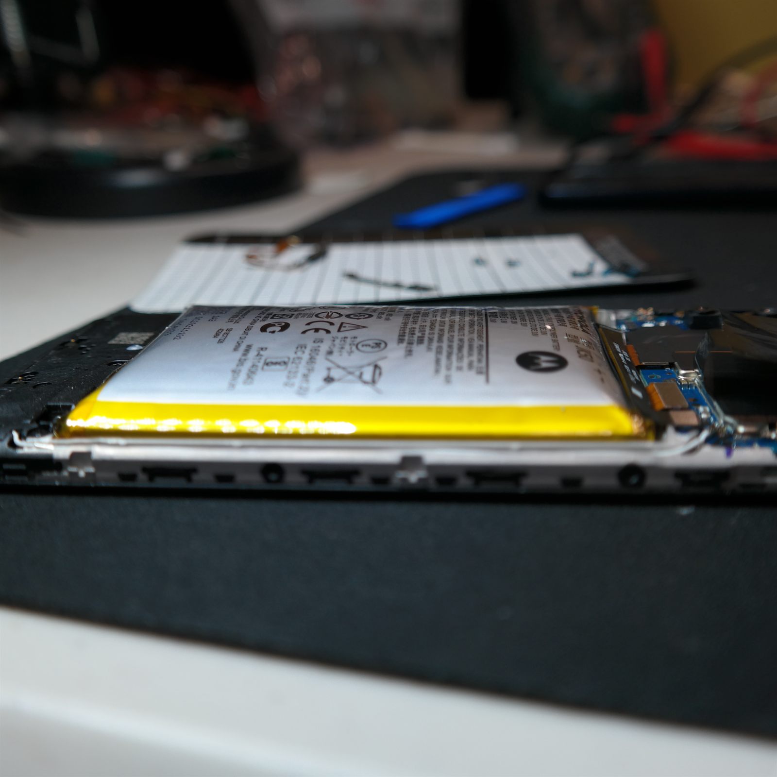 Low-angle close-up of the original battery still installed in the opened Moto G 5G (2022). The silver pouch cell bulges upward in a visible curve, standing proud of the metal chassis rail below it, with the Motorola logo and certification markings on its label.