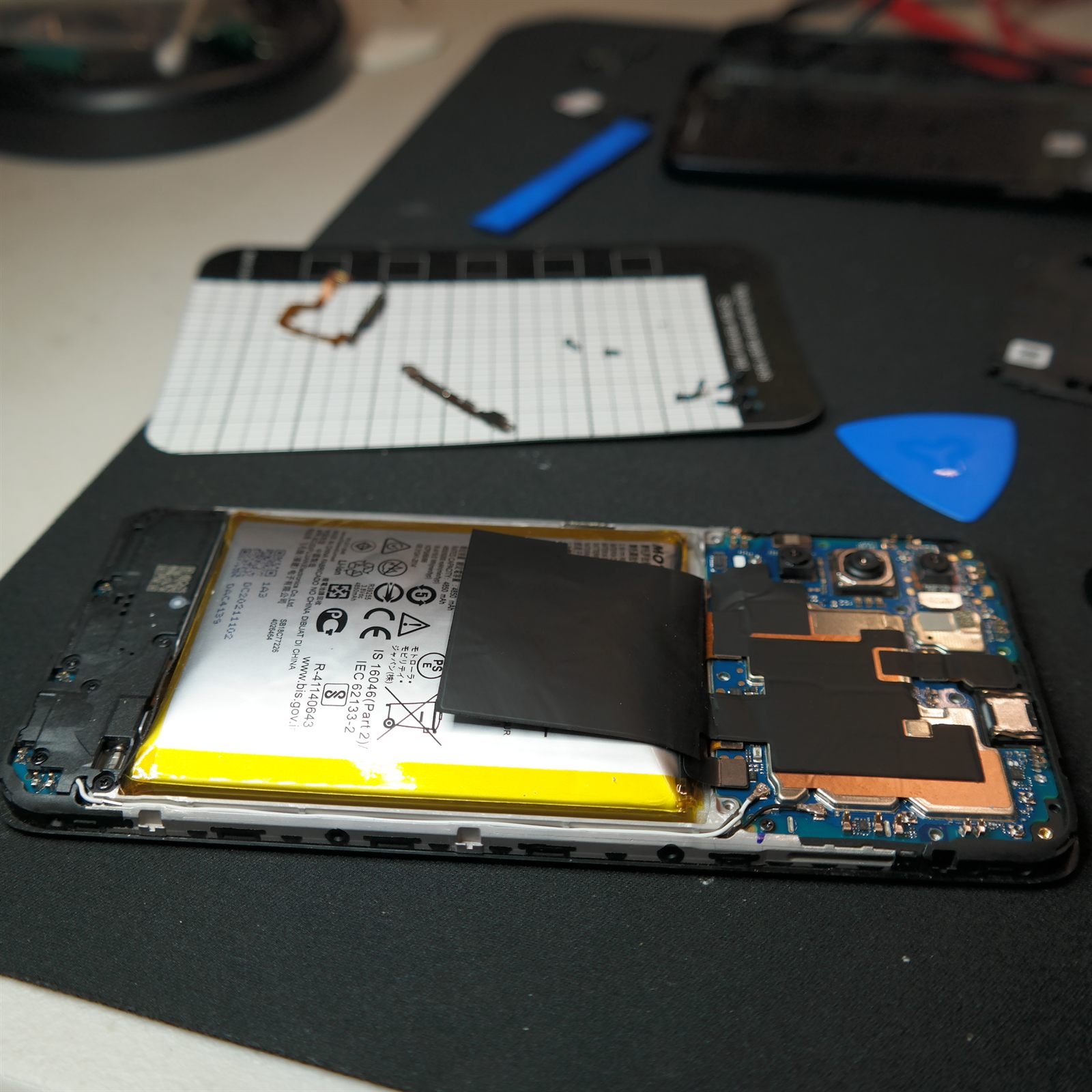 Interior of the Moto G 5G (2022) with the motherboard cover removed, exposing the swollen battery on the left and the main board with its copper shielding on the right. The removed power button, its bracket and its screws are laid out on a white magnetic parts mat behind the phone.