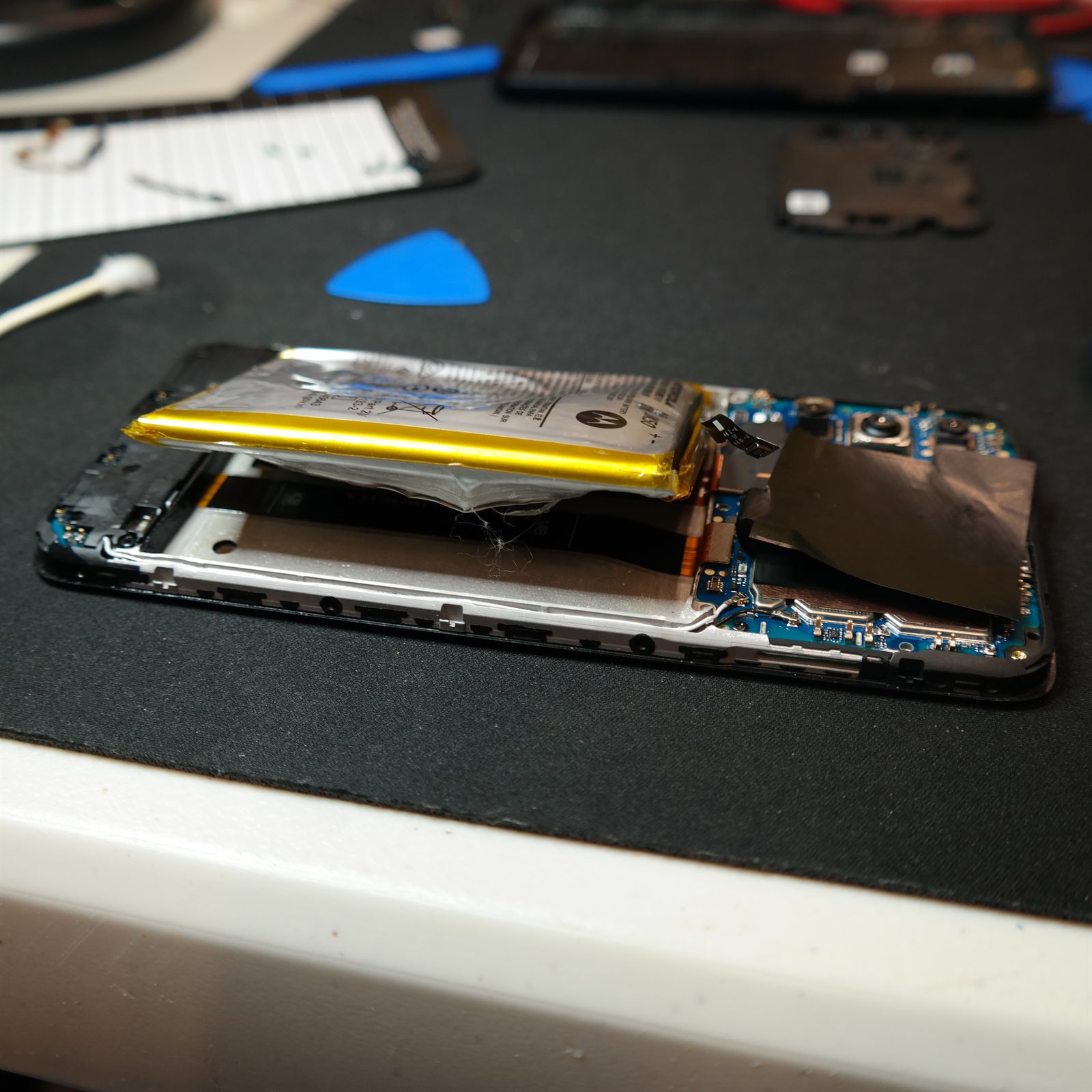 The swollen original battery lifted clear of the Moto G 5G (2022) chassis and resting on top of the open phone. The silver pouch is visibly wrinkled and distended rather than flat, with its yellow edge tape buckled.