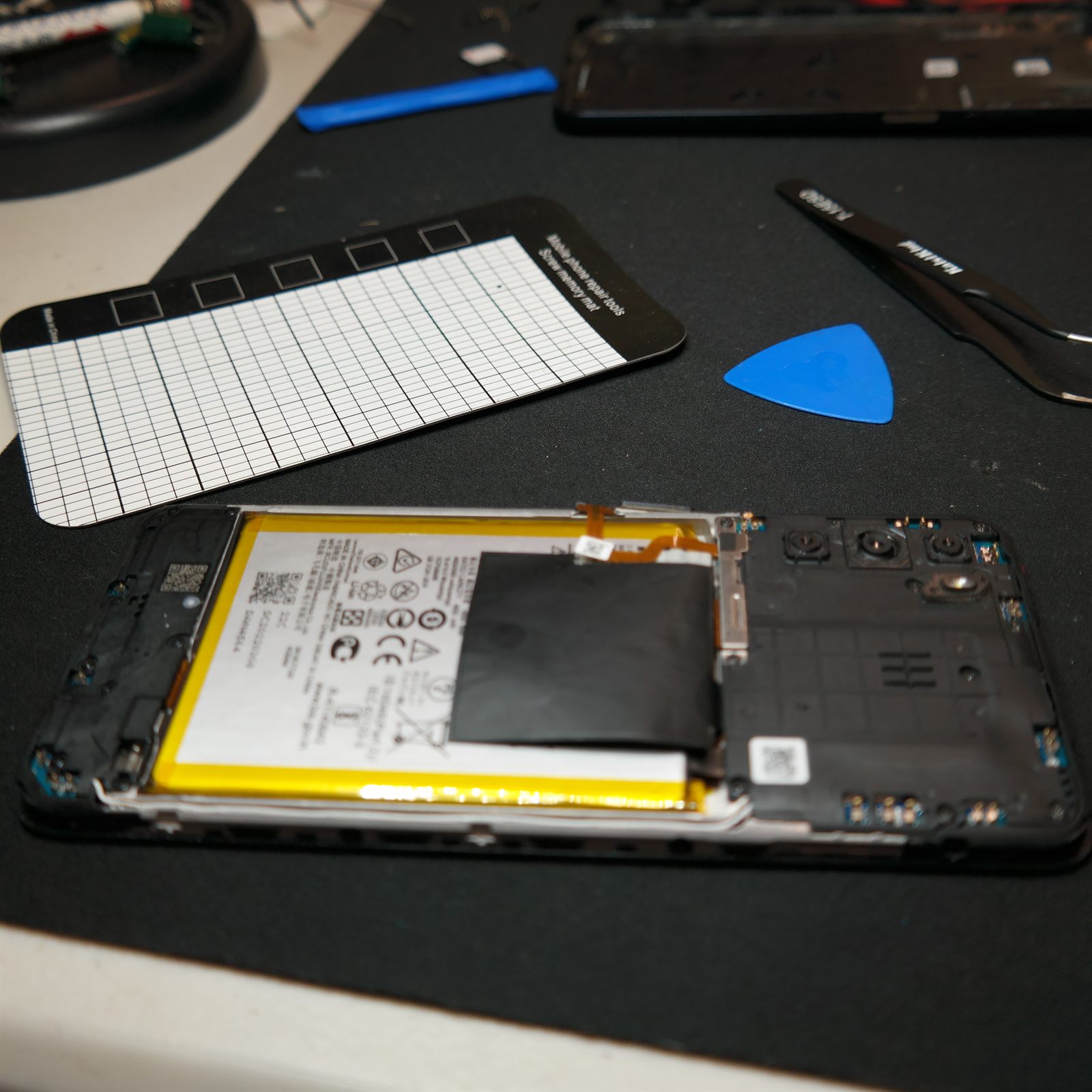 The flat replacement JK50 battery seated in the Moto G 5G (2022) chassis during reassembly, with the motherboard cover refitted to the right. A magnetic screw mat, tweezers and a blue opening pick sit on the bench above the phone.
