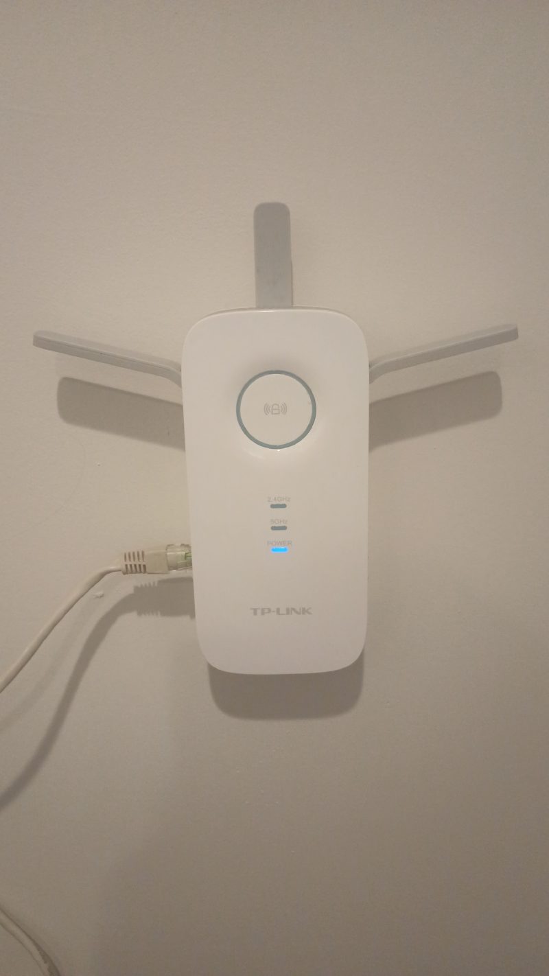 A white TP-Link range extender plugged into an outlet on a plain wall. Three antennas fan out from the top, one upright and two angled down to the sides. A white Ethernet cable with a translucent connector is plugged into the extender's left edge and runs down and out of the frame. A blue POWER indicator is lit below two unlit indicators labelled 2.4GHz and 5GHz.